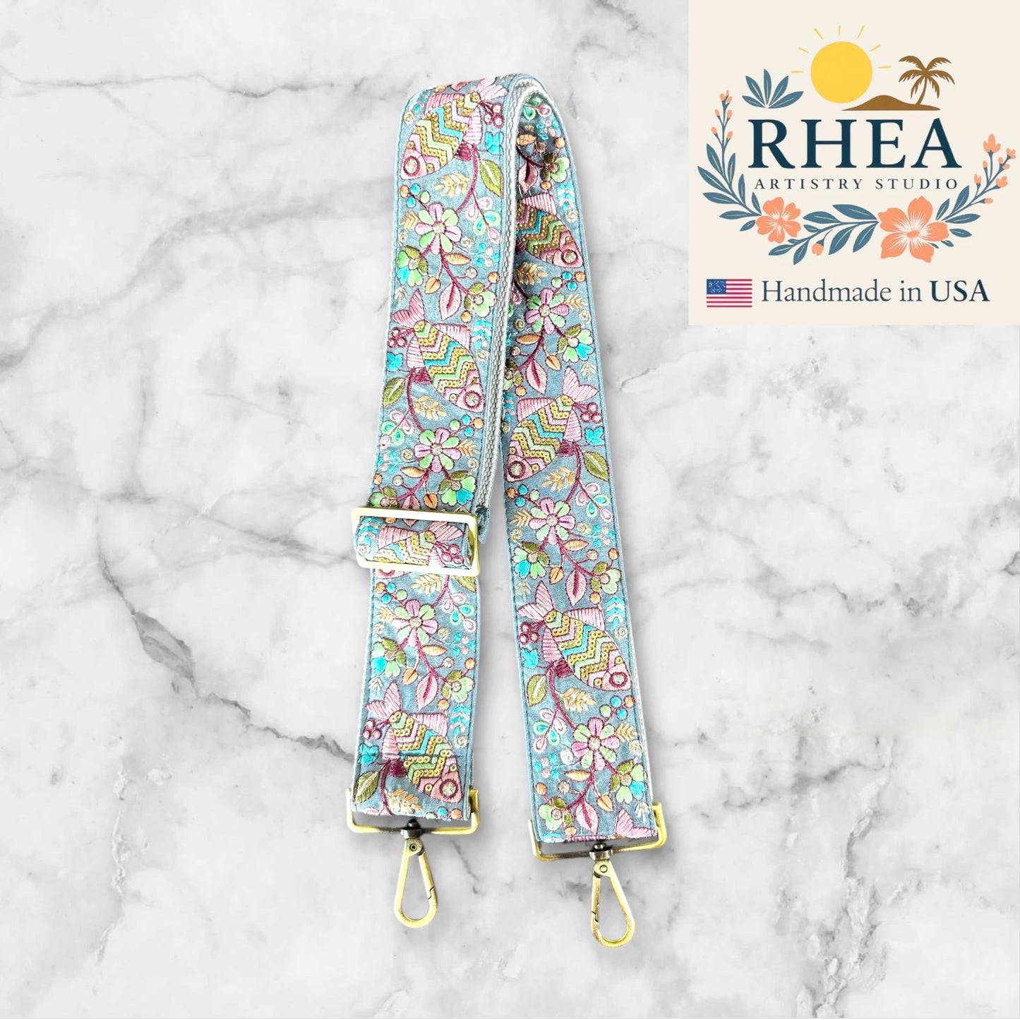 Floral patterned purse strap with gold hardware on a marble background, featuring RHEA Artistry Studio branding.