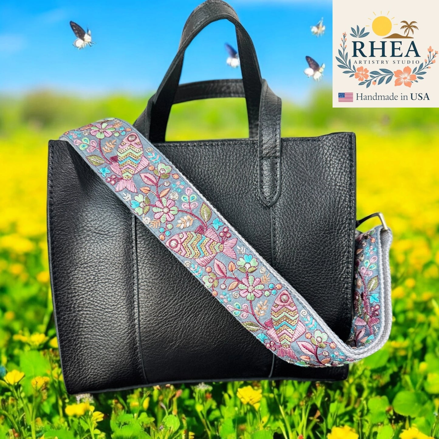 Black handbag with a colorful floral purse strap in a field with flowers and butterflies, featuring the RHEA brand.