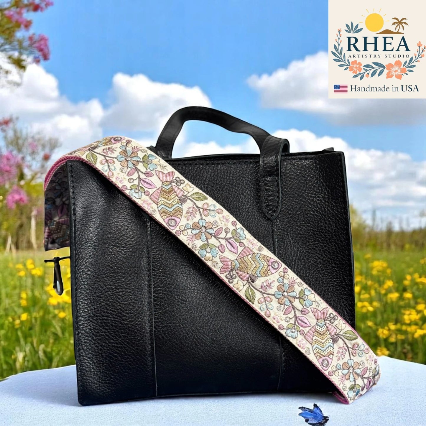 Black leather bag with a floral purse strap in a field with 'RHEA Artisan Studio' branding.