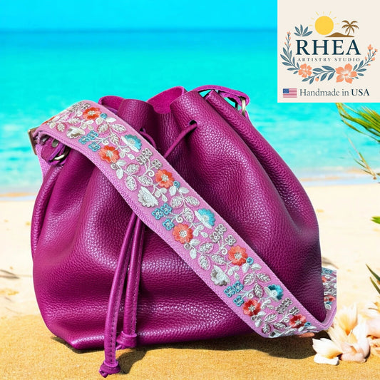 Purple drawstring bag with floral embroidery on a beach background, featuring the RHEA Artistry Studio logo.