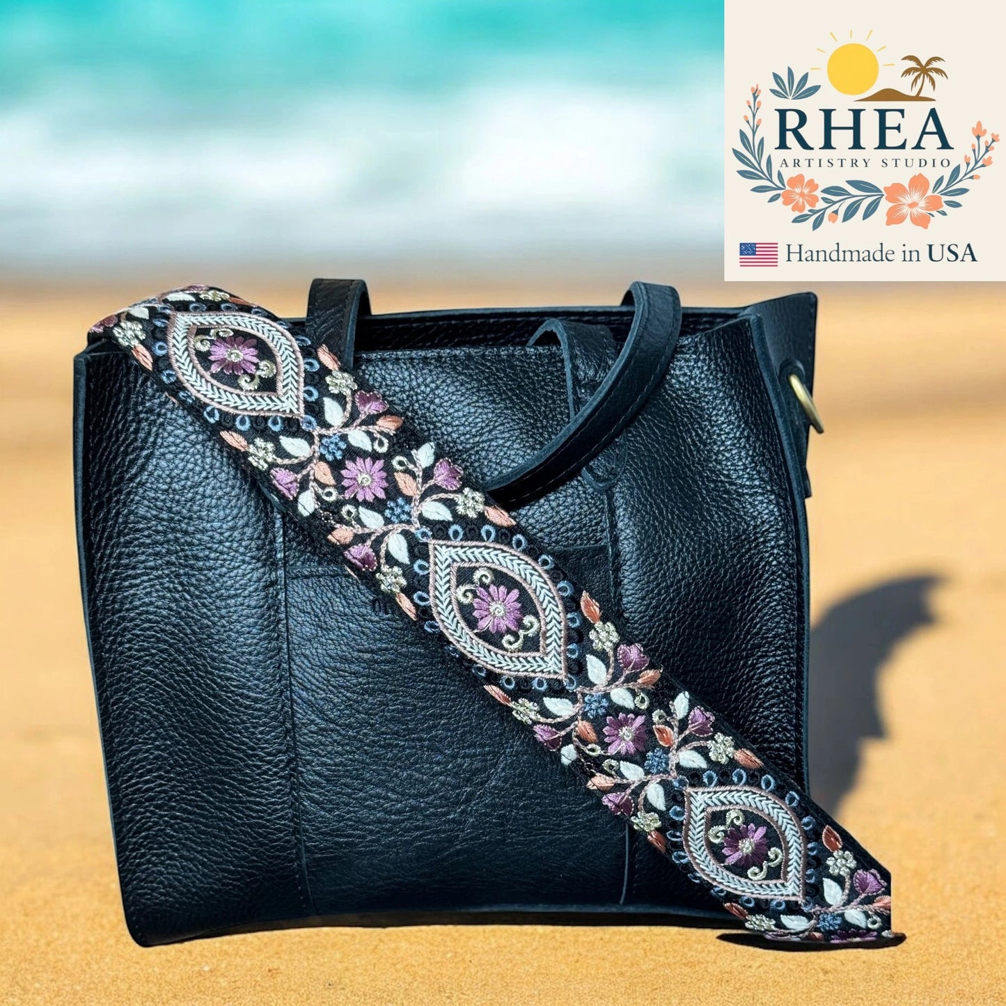 Black handbag with an ornate, colorful strap on a sandy background, featuring the RHEA Artistry Studio logo.