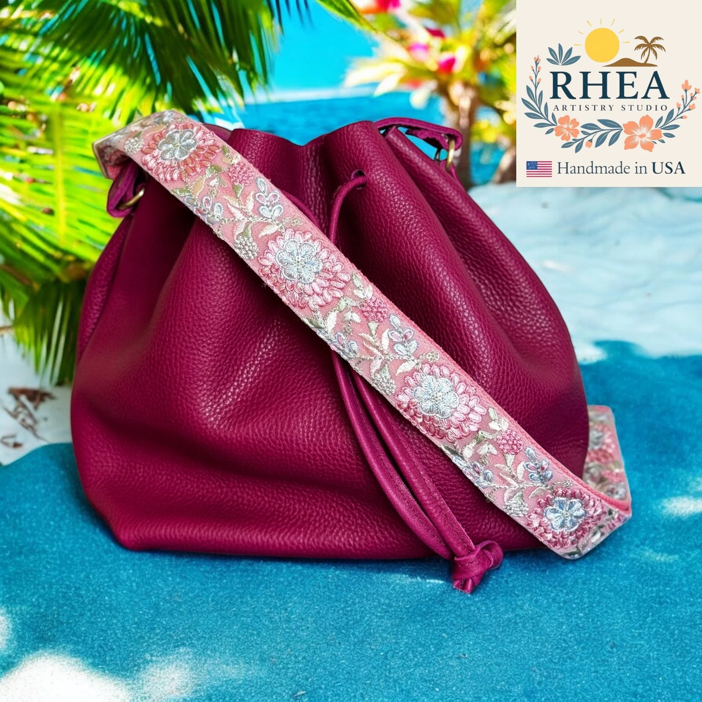 Pink leather bag with floral purse strap on a blue surface, RHEA Artistry Studio logo visible
