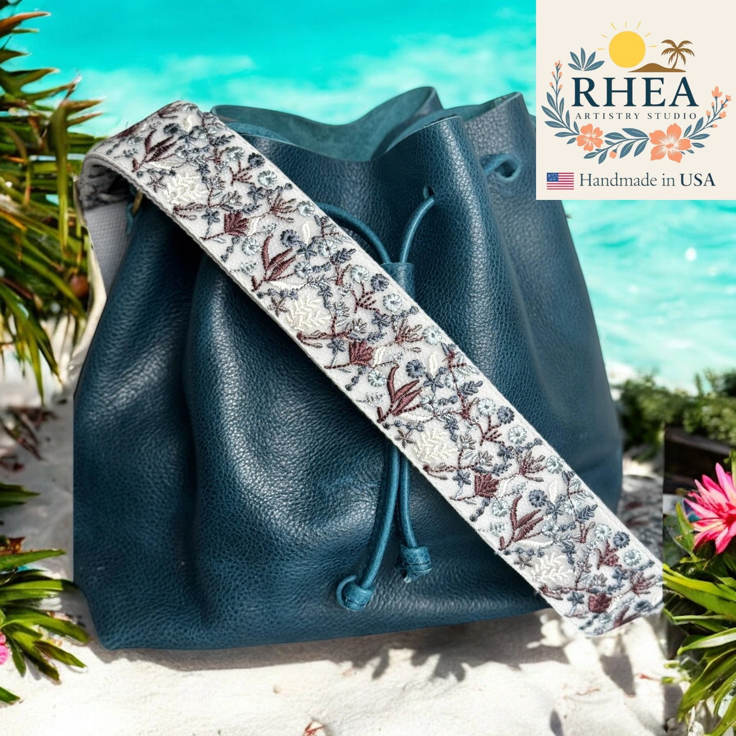 Blue leather bag with a floral purse strap against a scenic background, featuring the Rhea Artistry Studio logo.