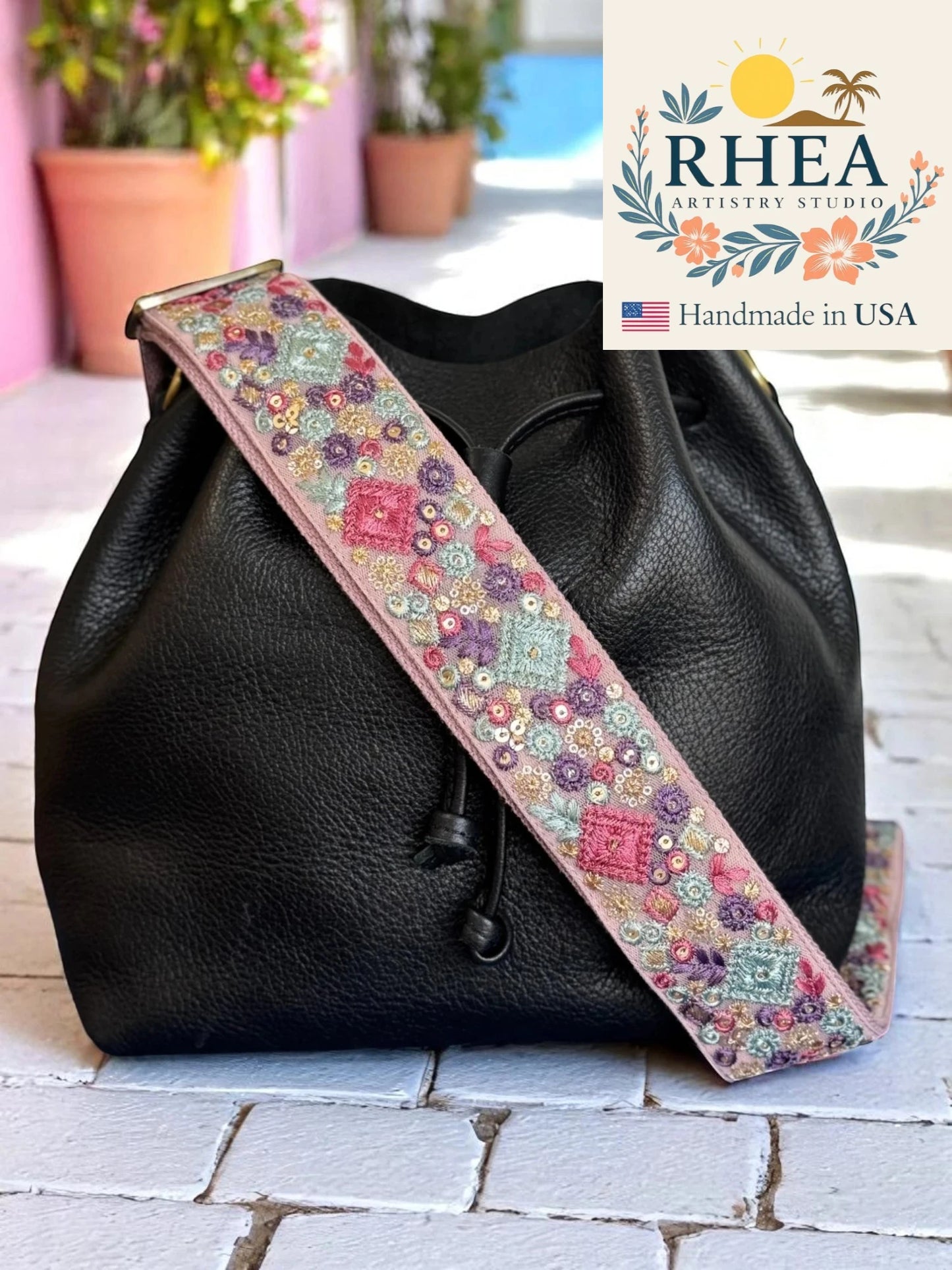 Black handbag with a colorful floral purse strap on a light wooden surface, featuring the RHEA Artistry Studio logo.