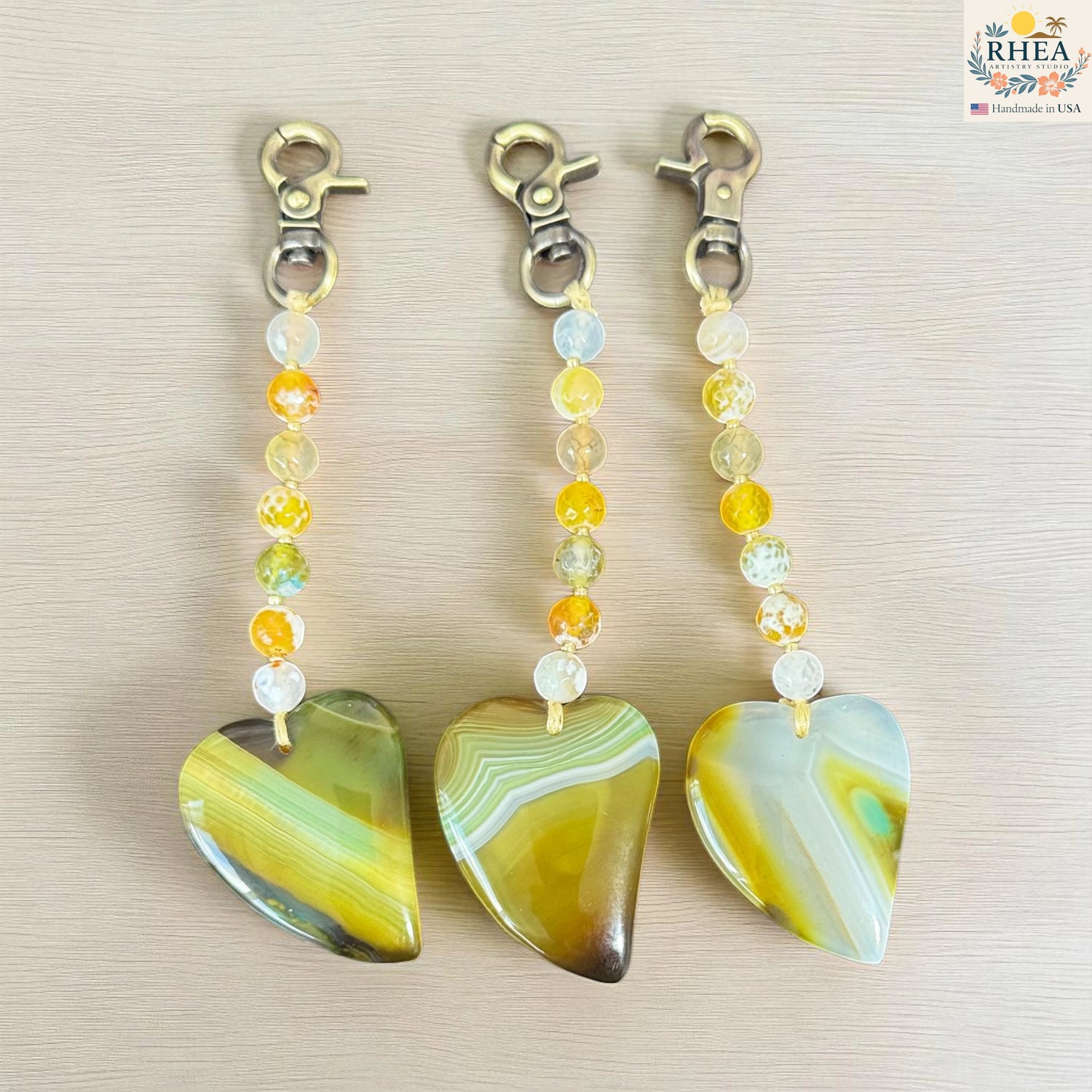 Three heart-shaped stone keychains with beaded extensions on a beige background, branded 'RHEA'.