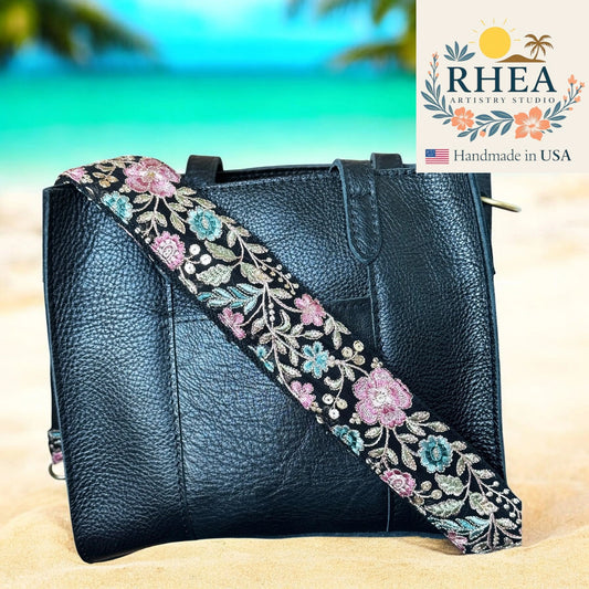 Black leather handbag with a floral embroidered strap on a beach background, featuring the RHEA Artistry Studio logo.