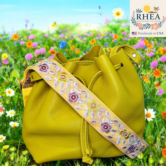 Yellow leather handbag with floral purse strap in a field of flowers, featuring the RHEA brand.