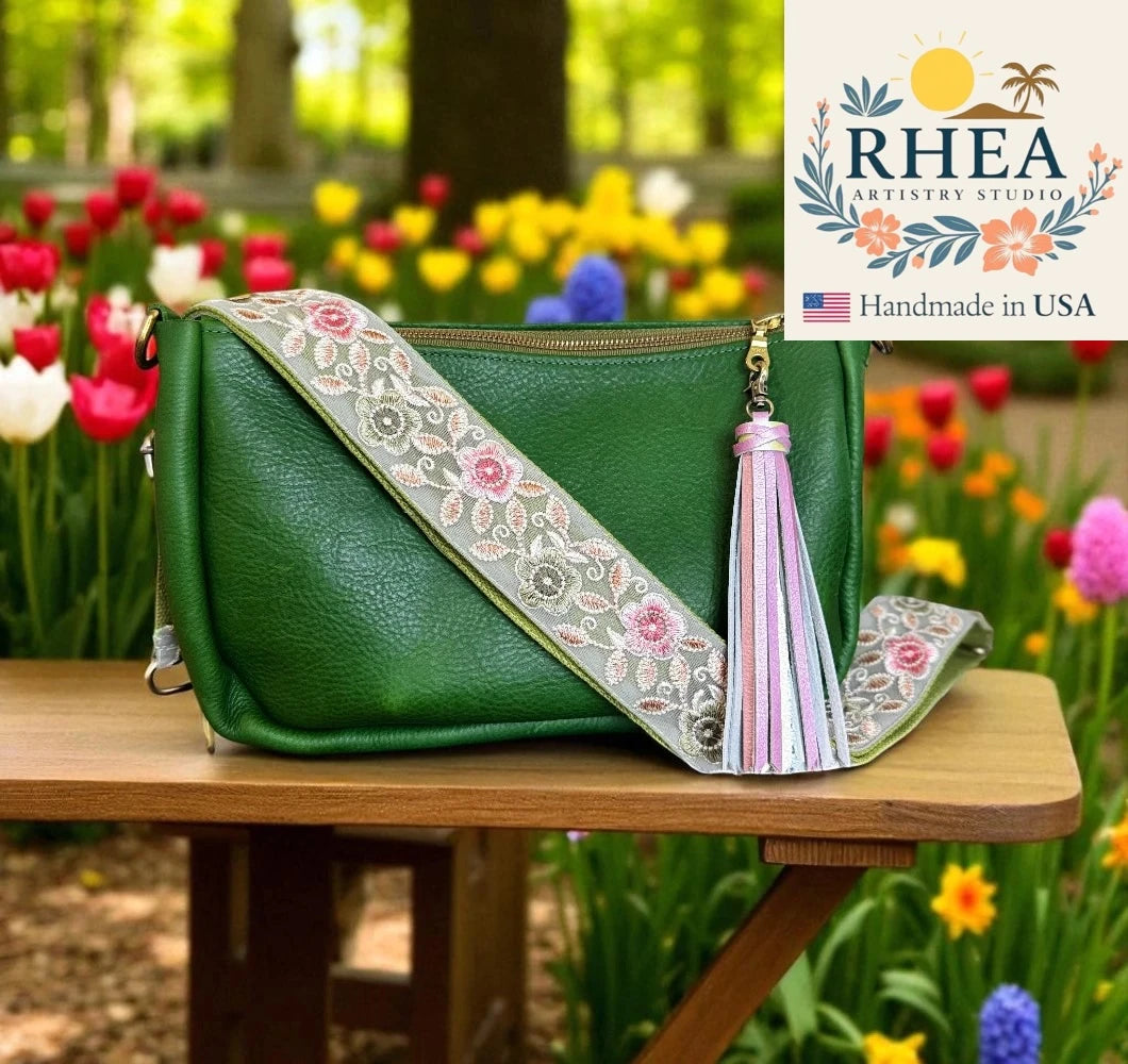 Green handbag with floral purse strap and tassel on a wooden table with a garden background, featuring RHEA Artistry Studio logo.