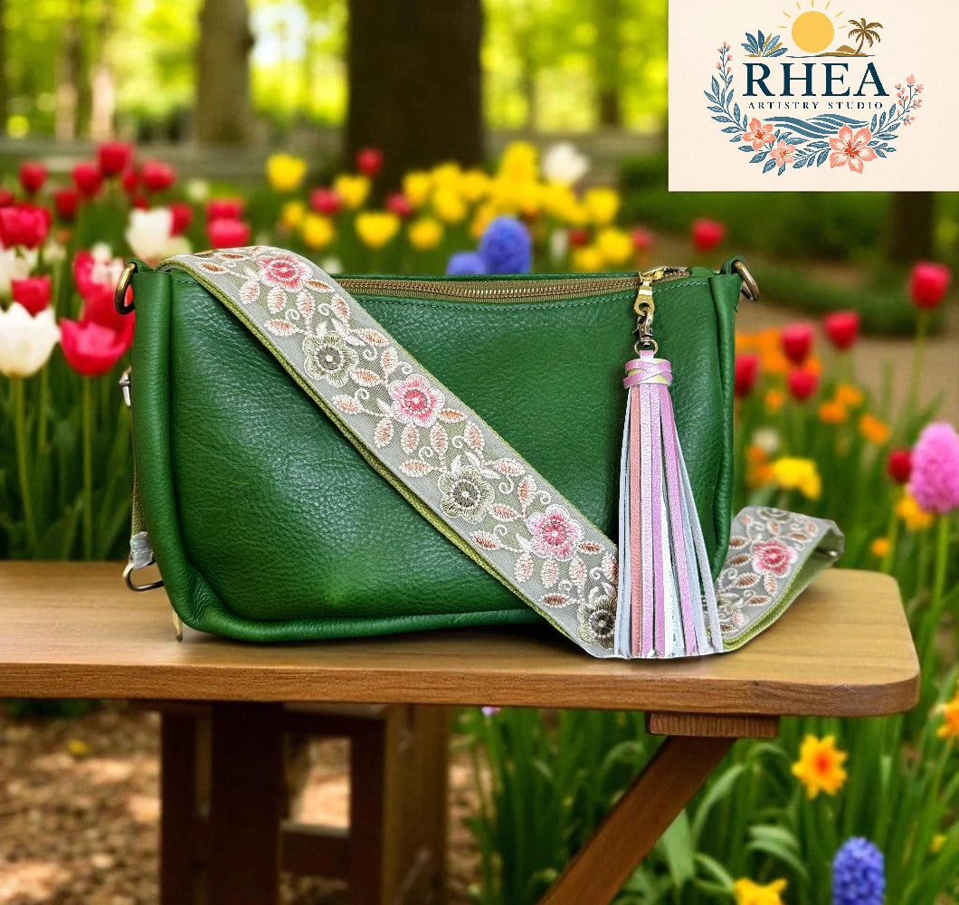Green handbag with floral strap and tassel on a wooden table with a garden background, featuring the RHEA brand.