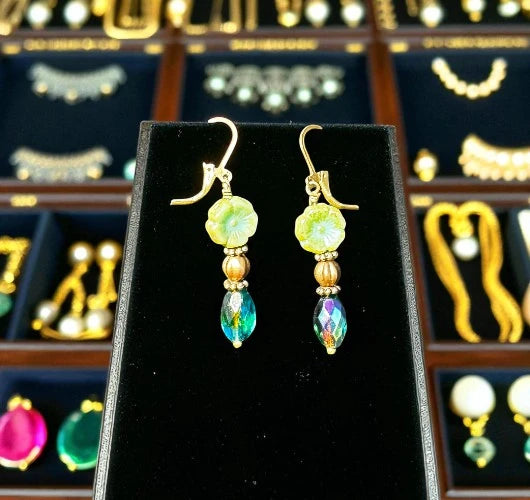 Gold and green beaded earrings on a black display stand with jewelry store background.