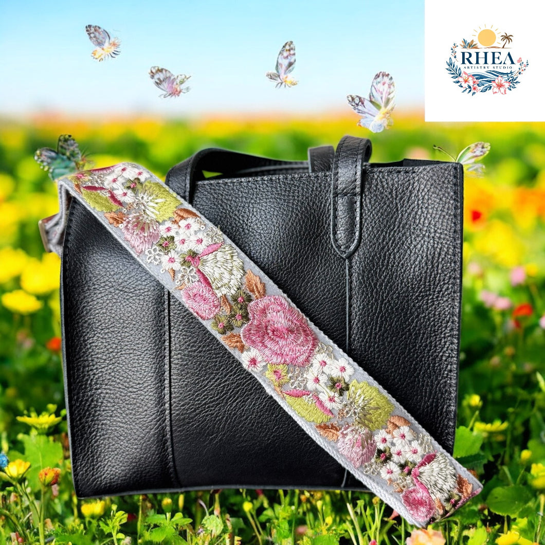 A black leather bag with a pastel strap embroidered in soft pinks, green, and white, photographed in a blooming field of wildflowers.