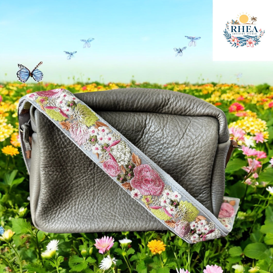 A taupe leather bag with a pastel strap embroidered in soft pinks, green, and white, photographed in a blooming field of wildflowers.