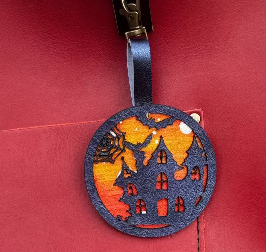 A handpainted leather charm with a halloweendesign, attached to a red leather bag.