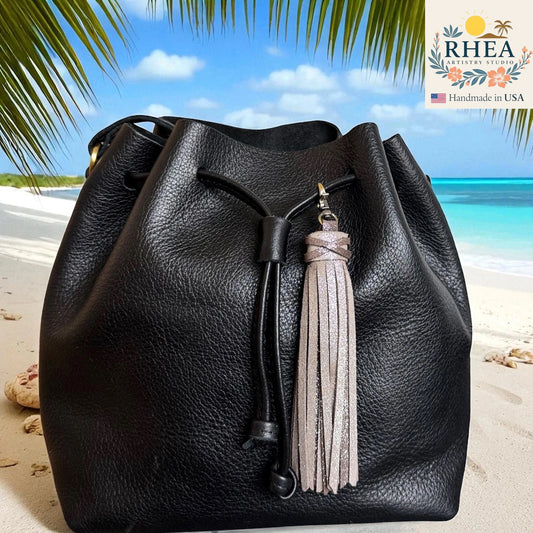 Black leather bucket bag with a tassel on a beach background, featuring the RHEA brand.