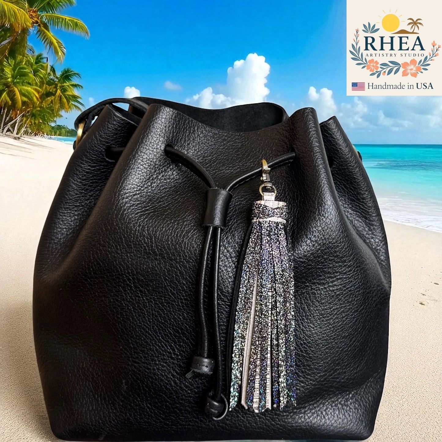 Black leather bucket bag with a tassel on a beach background, featuring the RHEEA brand.