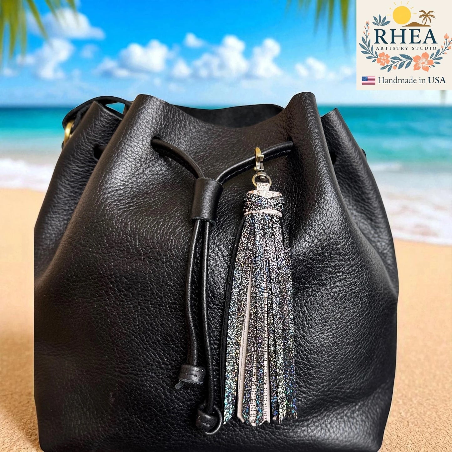 Black leather drawstring bag with a tassel on a beach background, featuring the RHEA brand.