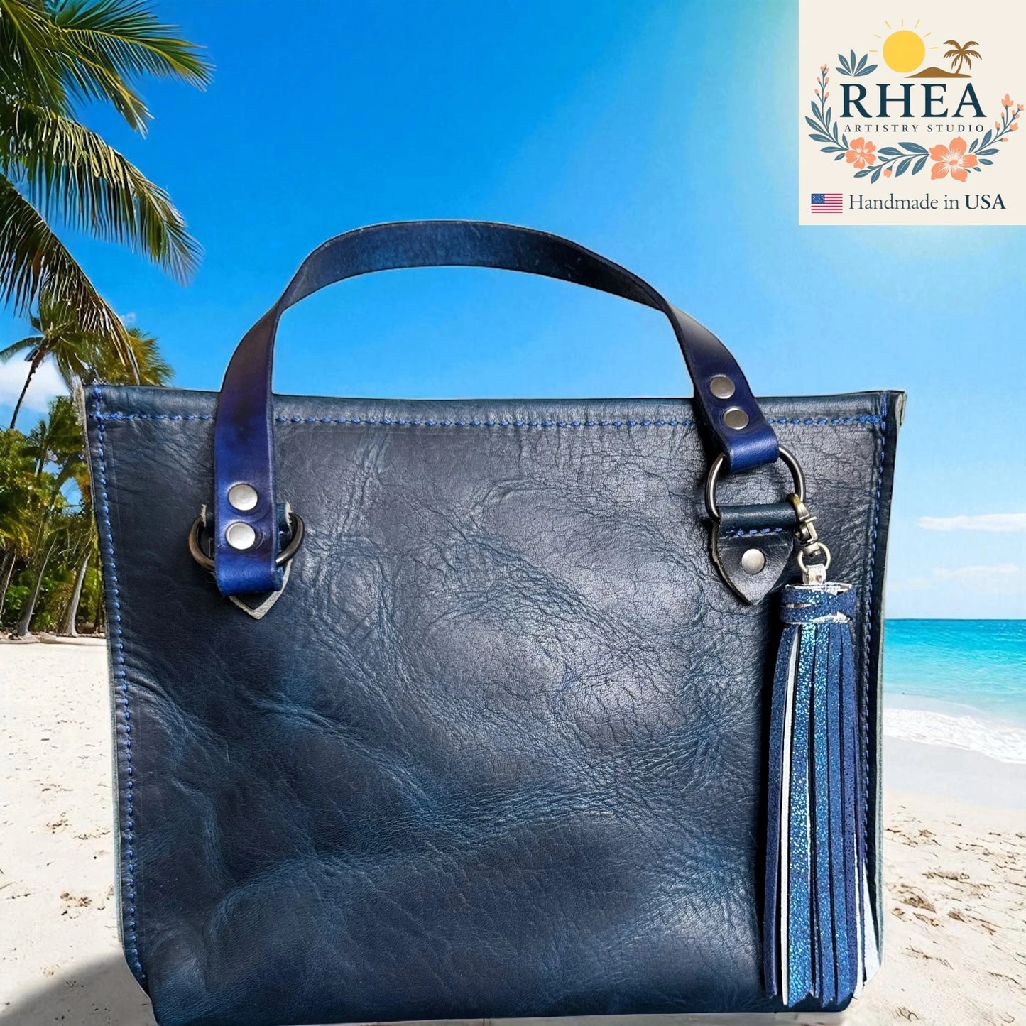 Blue leather handbag with tassel on a beach, featuring RHEA branding.