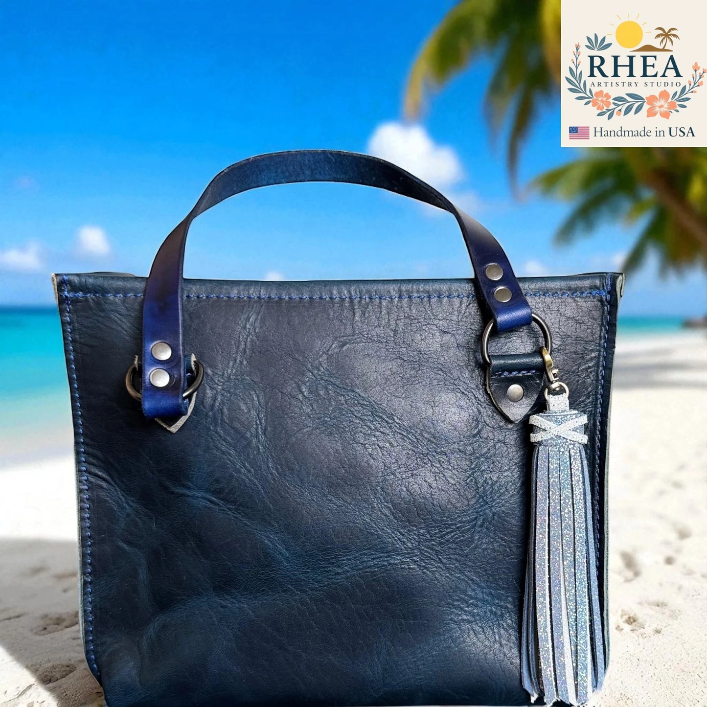 Blue leather handbag with tassel on a beach, featuring the RHEA Artistry Studio logo.