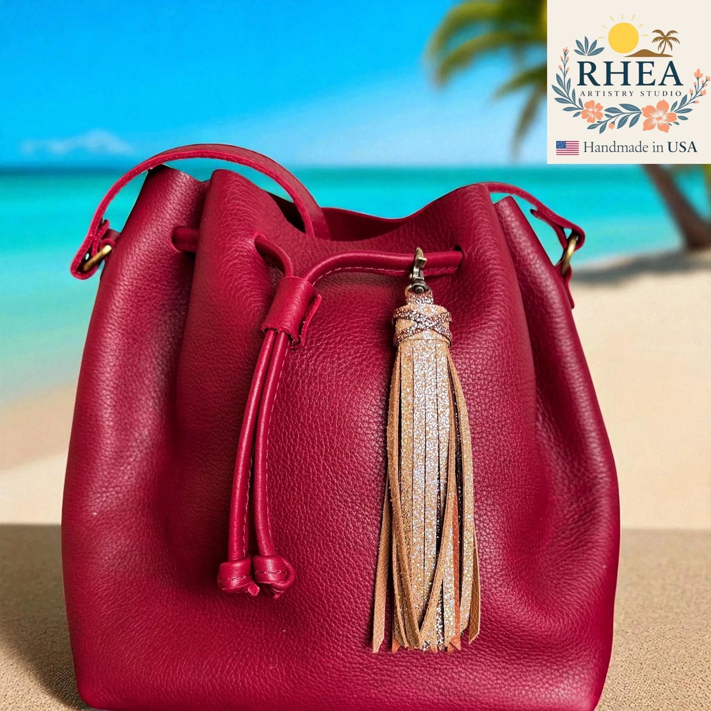 Red leather bucket bag with a tassel on a beach background, featuring the RHEA Artisan Studio logo.