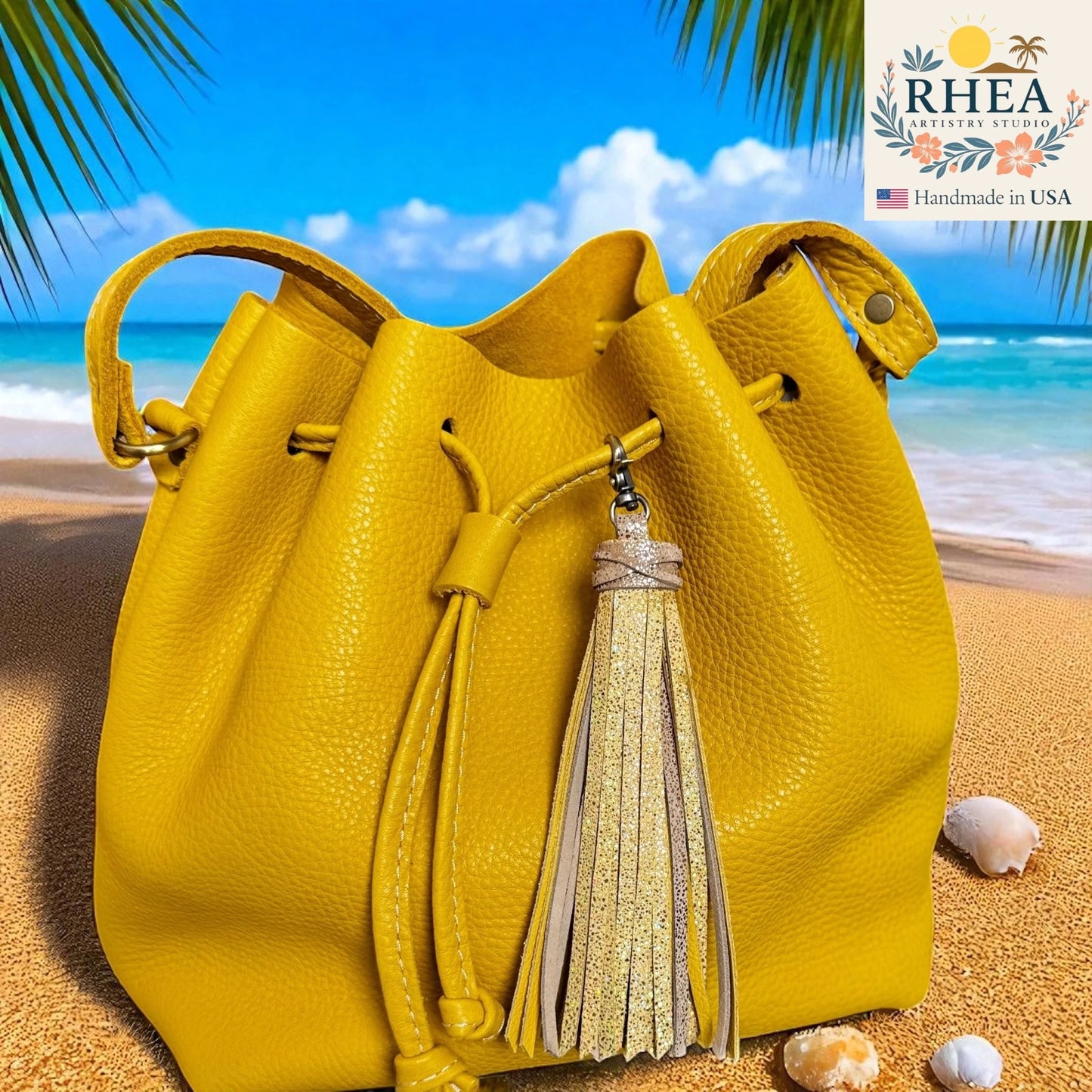 Yellow leather bucket bag with tassel on a beach, featuring the RHEA brand.