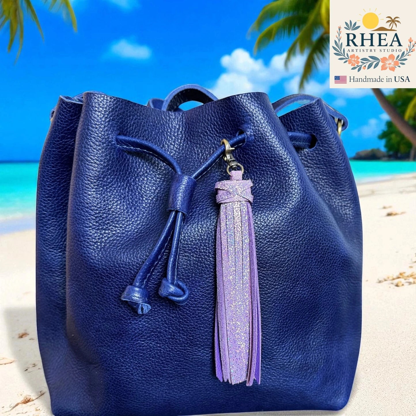 Blue leather bag with a purple tassel on a beach background, featuring the RHEA Artistry Studio logo.