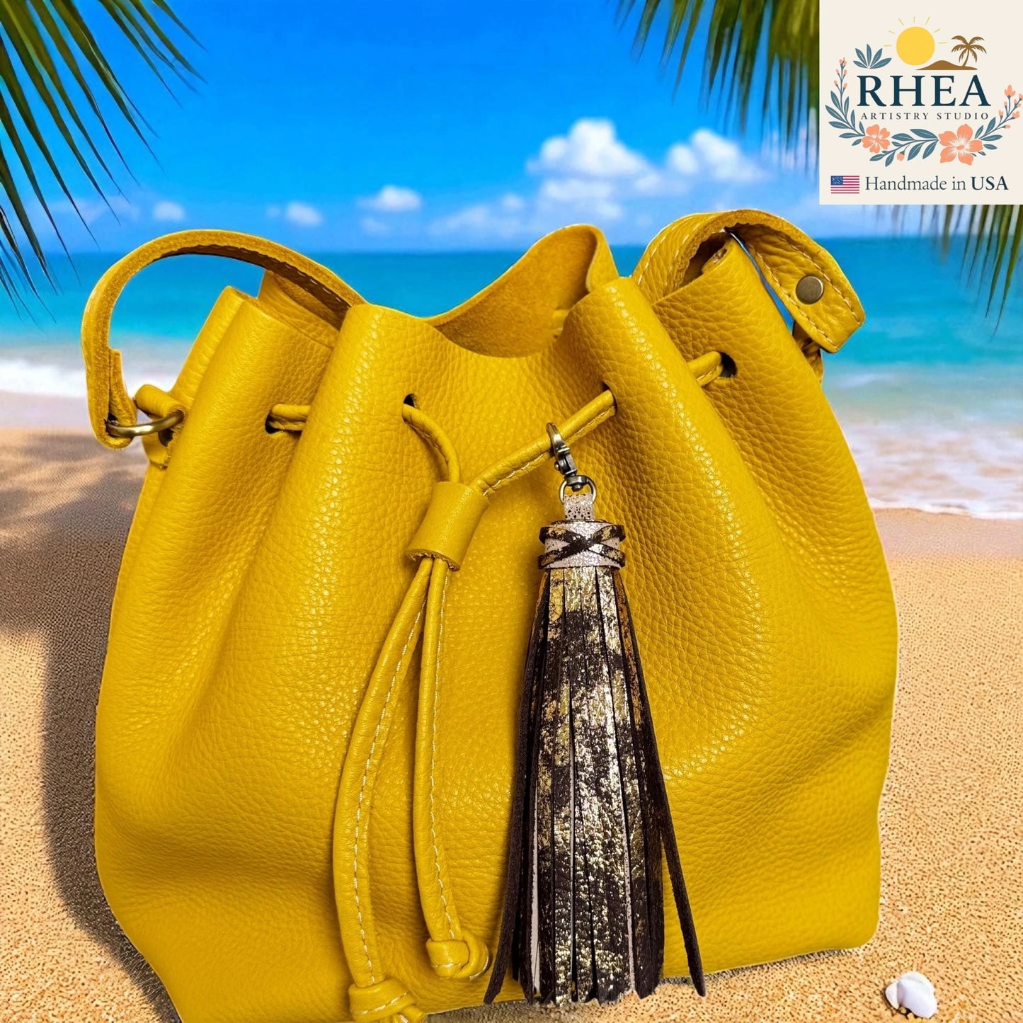 Yellow leather bag with a tassel on a beach background, featuring the RHEA brand.