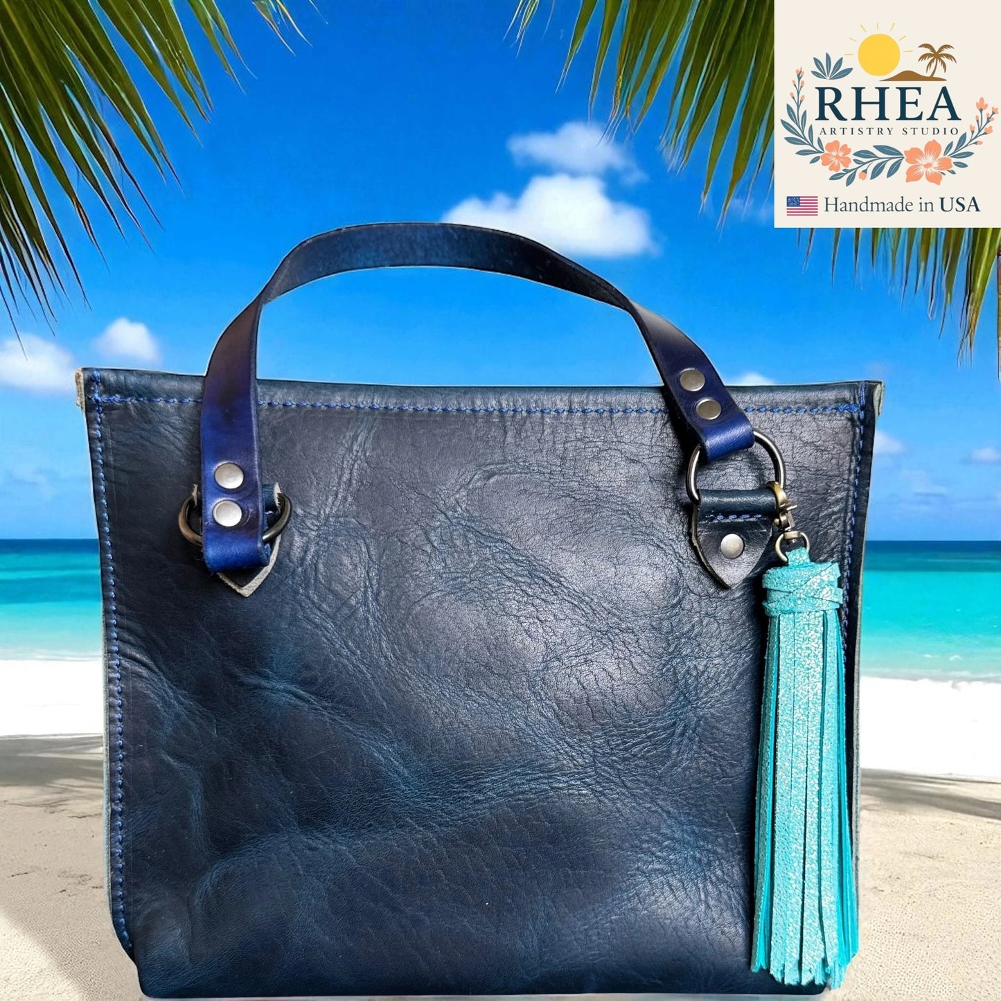 Blue leather handbag with a tassel on a beach background, featuring the RHEA Artisan Studio logo.