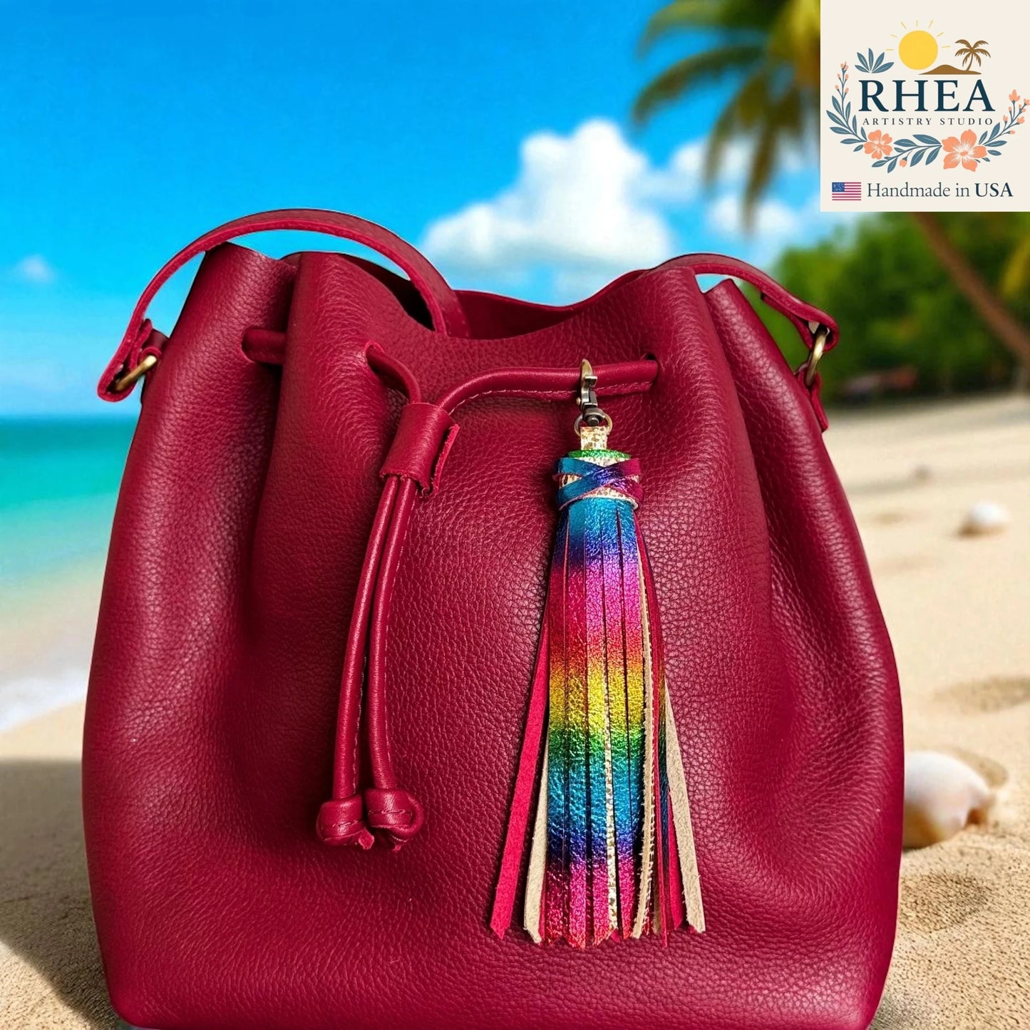 Red leather bucket bag with a colorful tassel on a beach background, featuring the RHEA brand.