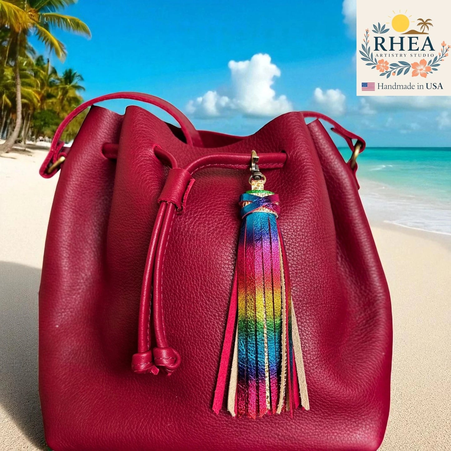 Red leather bucket bag with a colorful tassel on a beach background, featuring the RHEA brand.