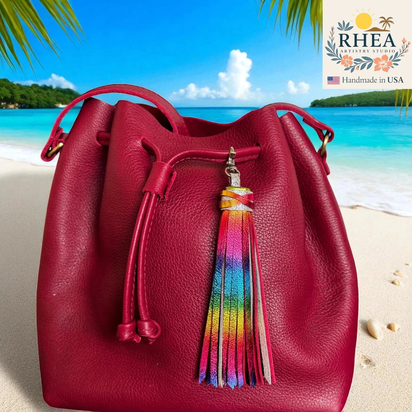 Red leather bucket bag with a colorful tassel on a beach background, featuring the RHEA brand.