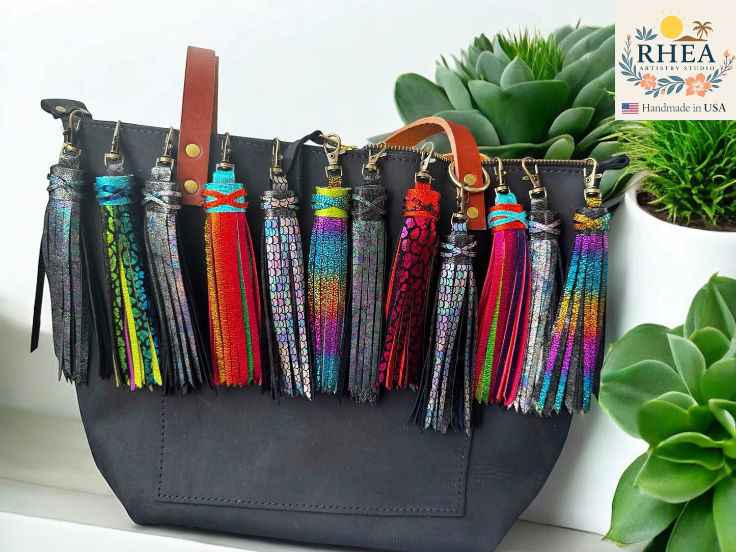 Black handbag with colorful tassels on a white surface with plants in the background, featuring the RHEA brand.