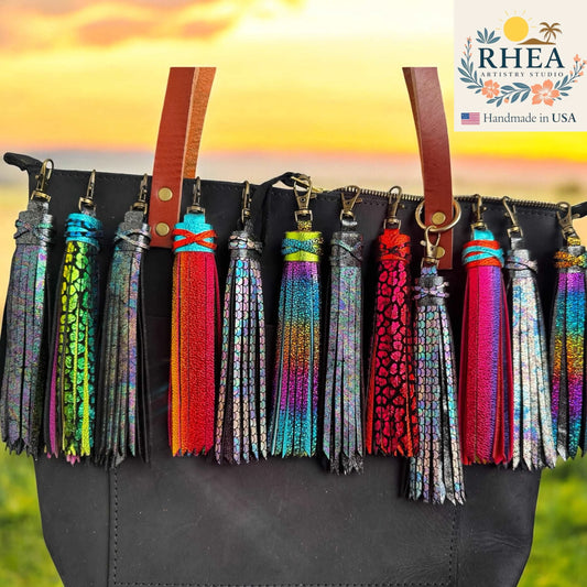 Colorful tassels on a black bag with a sunset background and RHEA branding.