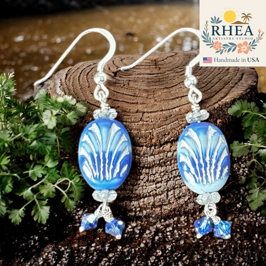 Blue and white patterned earrings on a wooden log with greenery, RHEA Artistry Studio logo visible.