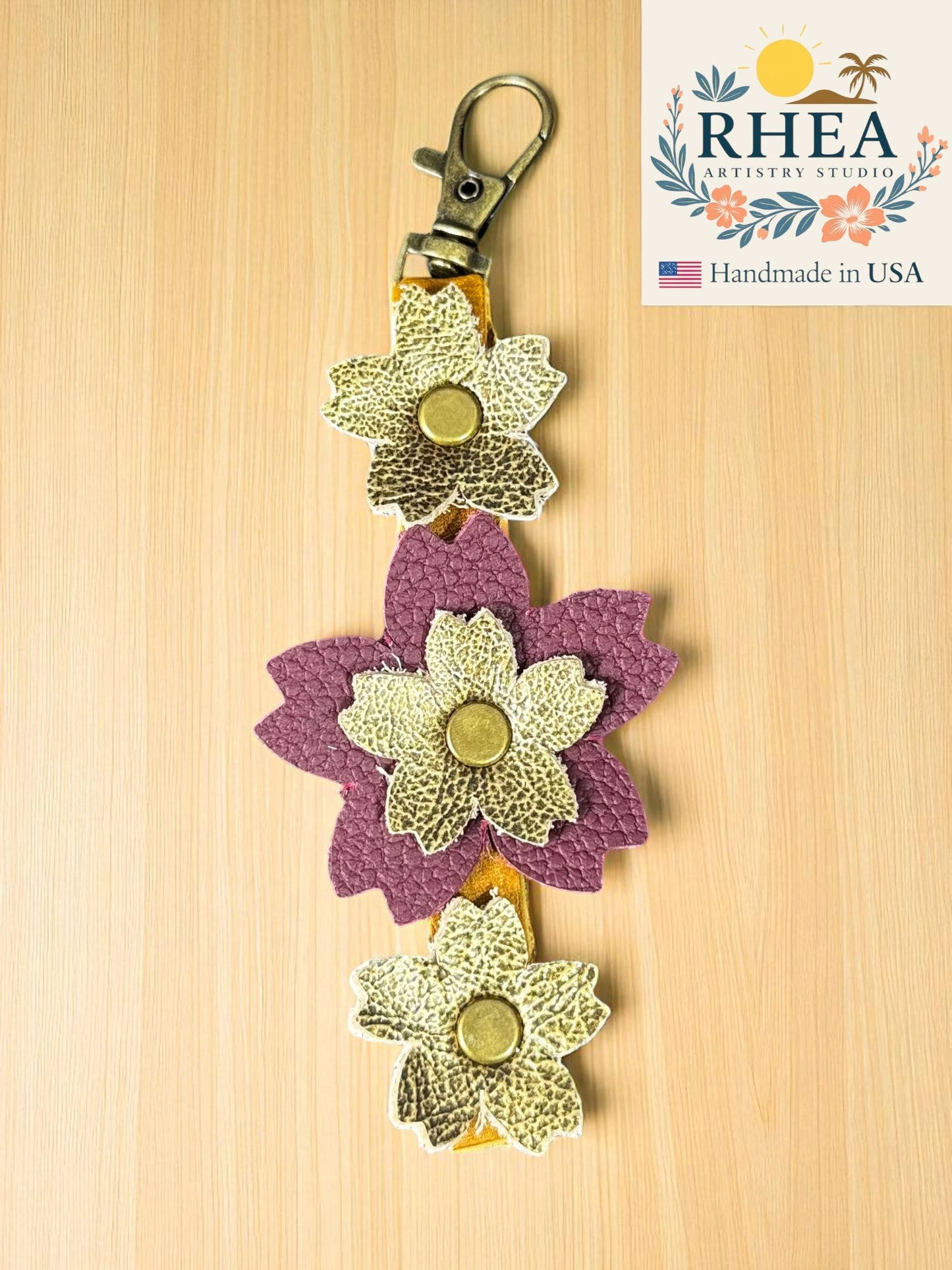 handmade leather bag charm with three flowers on a wooden background, featuring the RHEA brand.