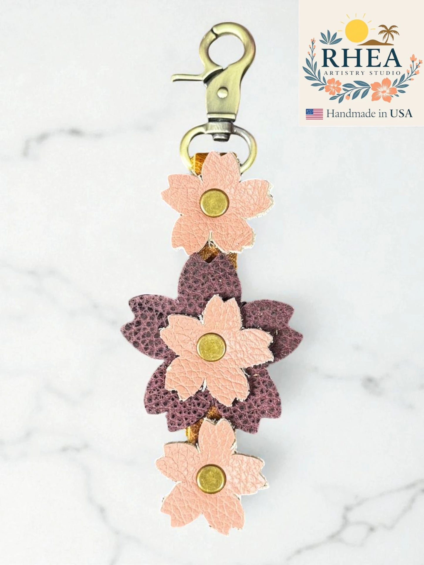 handmade leather bag charm with peach burgundy on a marble background, featuring RHEA Artisan Studio branding.
