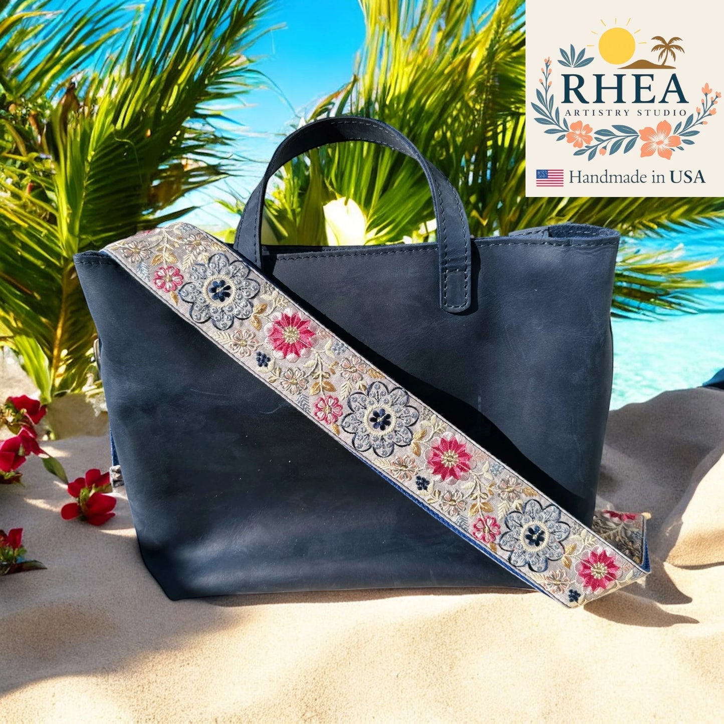 Navy blue handbag with floral purse strap on a beach background, featuring the RHEA Artistry Studio logo.