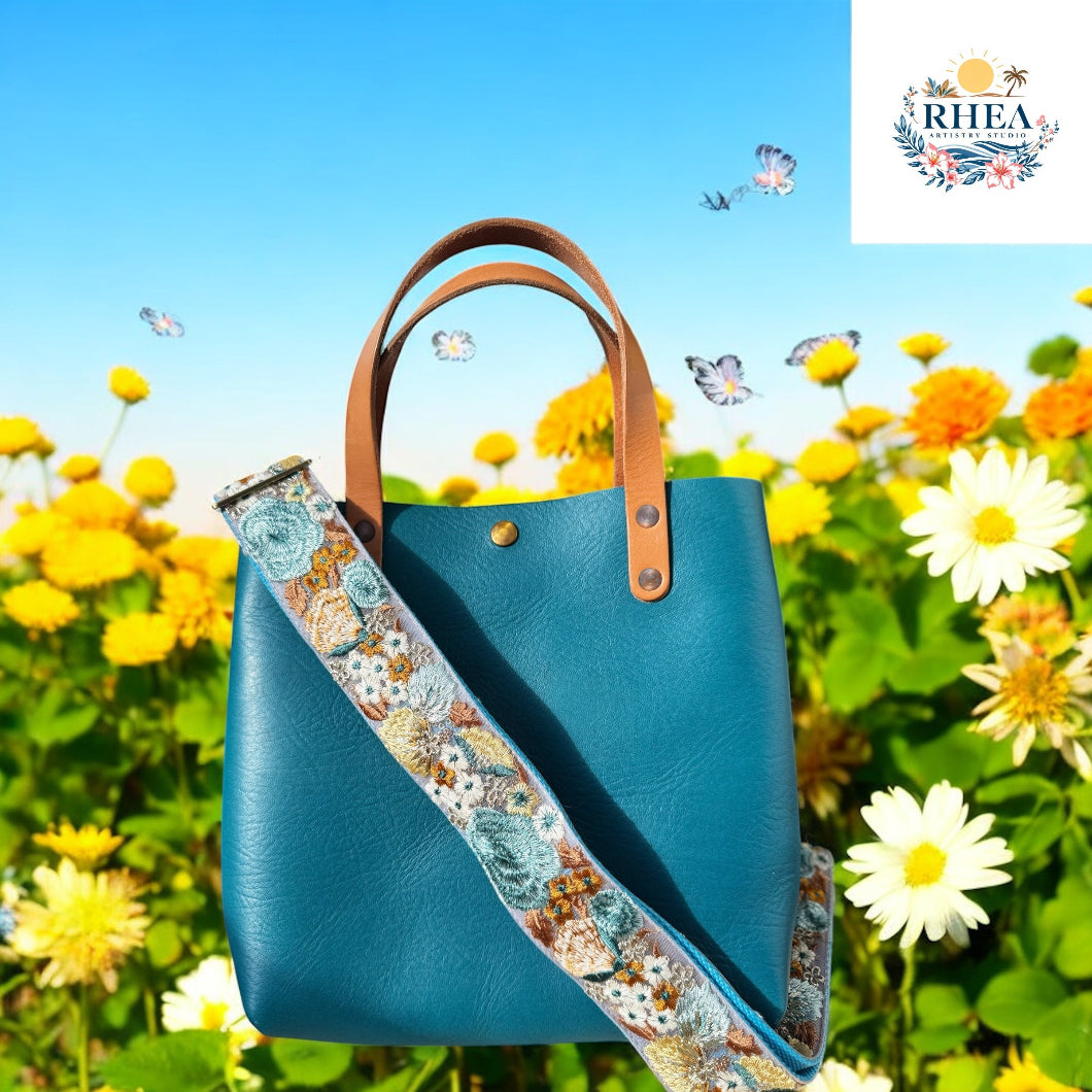 A teal handbag with natural tan handles and an intricately embroidered strap in dusty blue, ochre, and soft ivory florals, nestled in a radiant field of golden blooms under a cloudless sky.