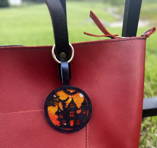 A handpainted leather charm with a halloweendesign, attached to a red leather bag.