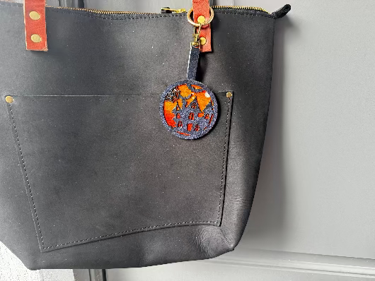 A handpainted leather charm with a halloweendesign, attached to a black leather bag.
