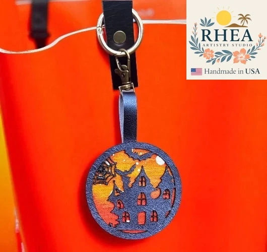 Decorative leather bag charm with house design on a red background, featuring RHEA Artisans Studio logo.