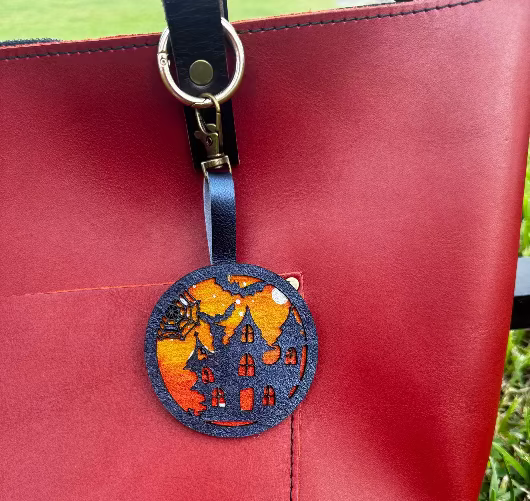 A handpainted leather charm with a halloweendesign, attached to a black leather bag.