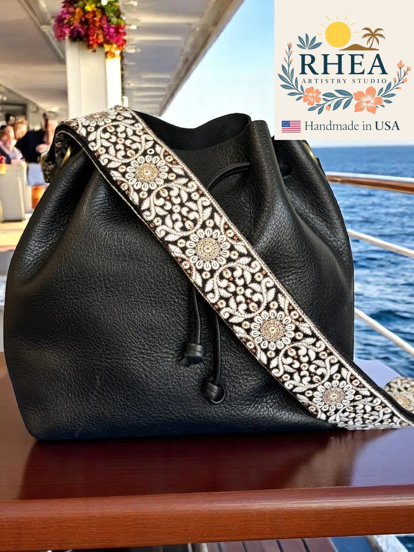Black leather handbag with decorative purse strap on a boat, featuring Rhea Artistry Studio branding.