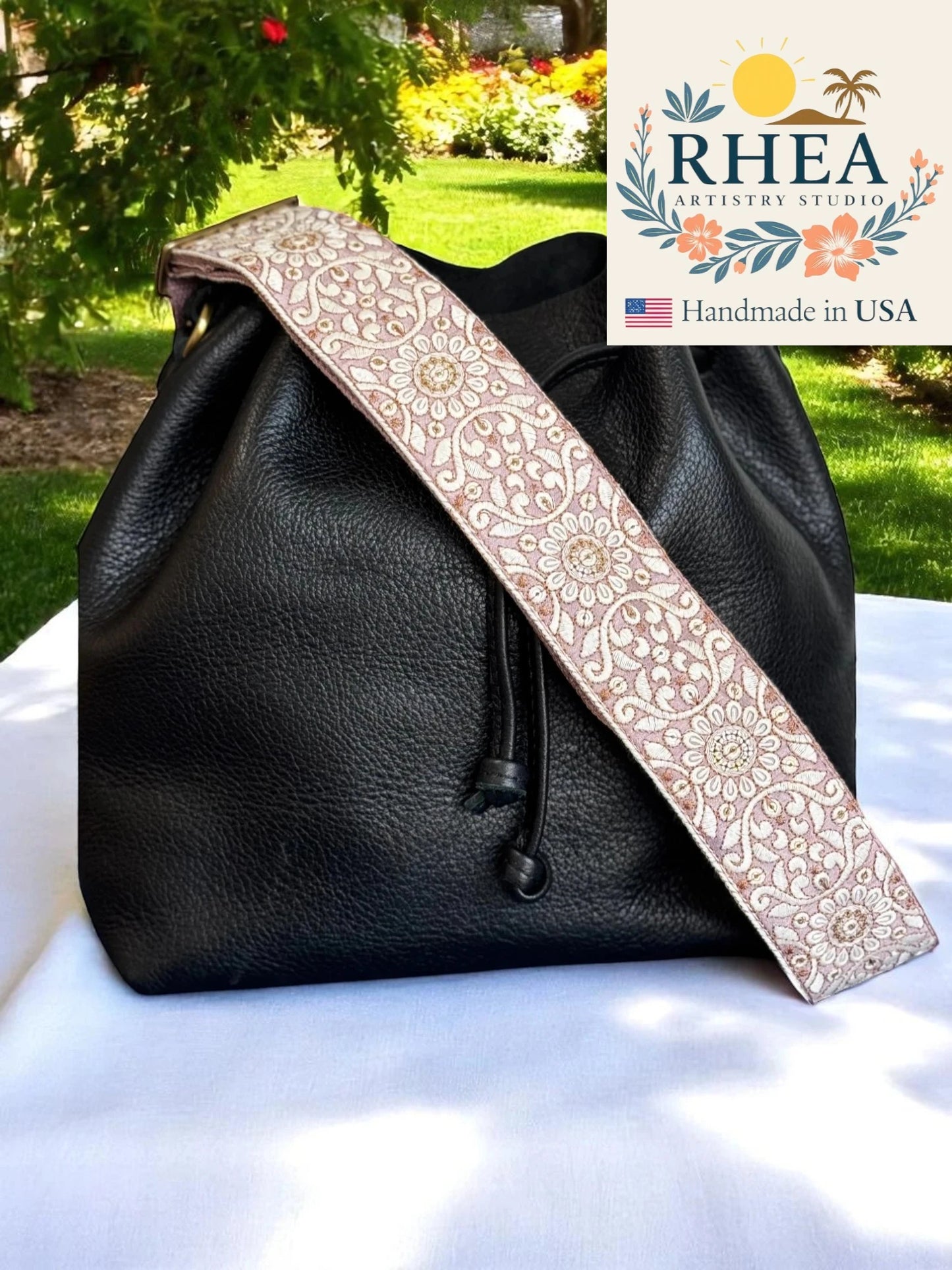 Black leather bag with a decorative purse strap on a white surface outdoors, featuring the RHEA Artisan Studio logo.