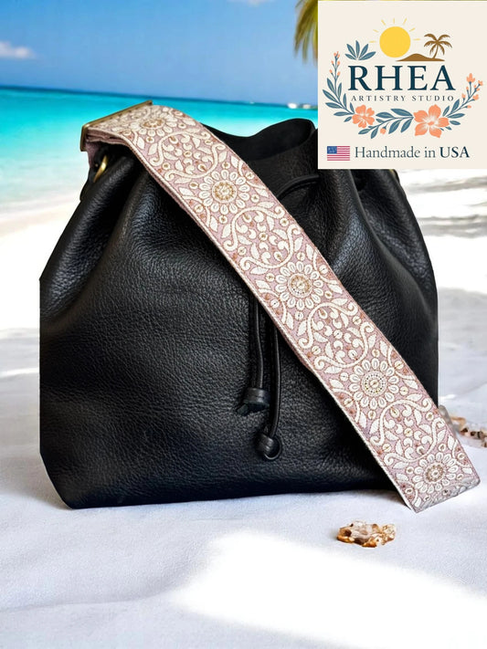 Black leather bag with a decorative purse strap on a sandy beach, featuring the Rhea Artistry Studio logo.