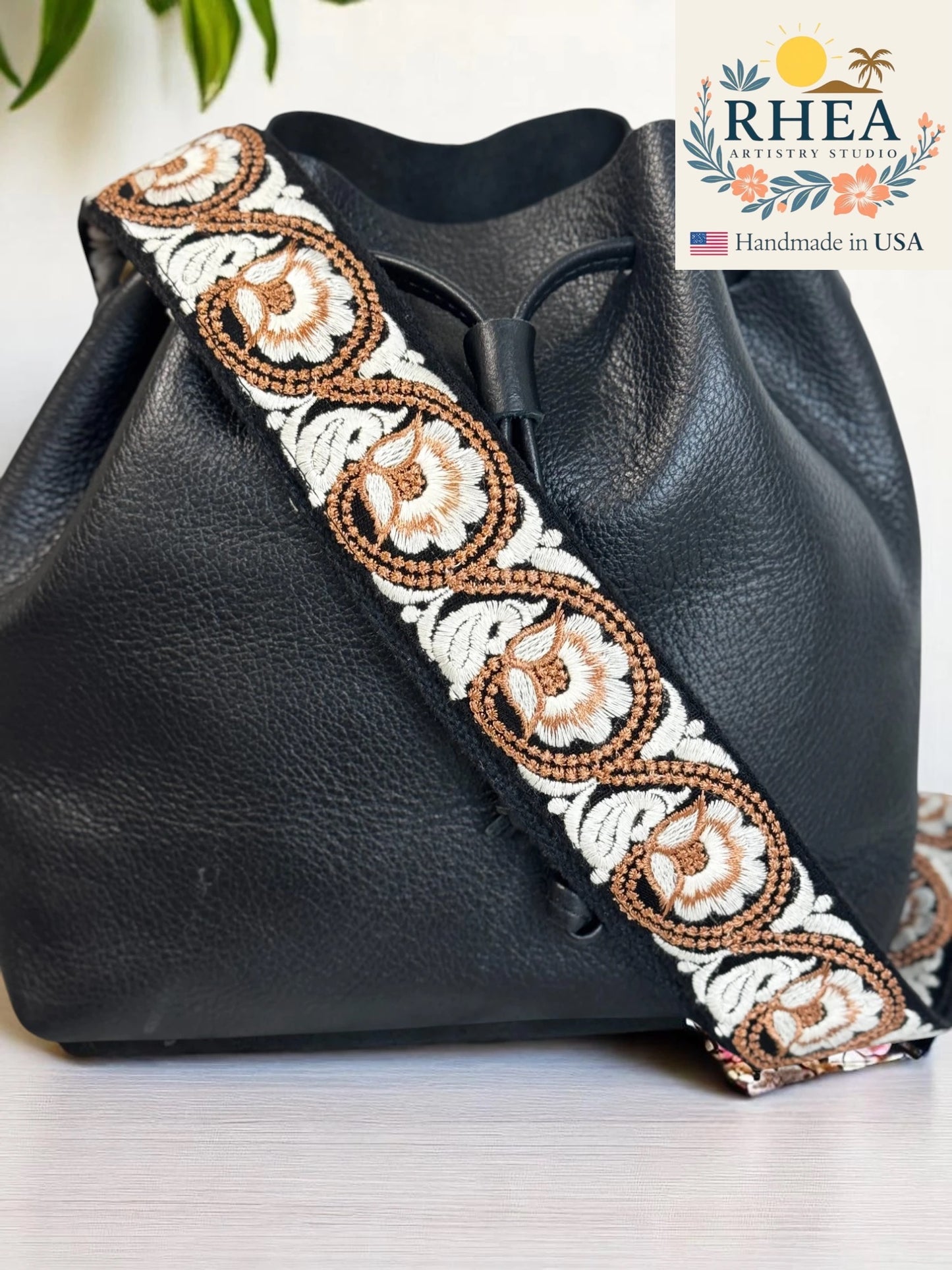Black leather bag with decorative purse strap featuring RHEA Artisans Studio branding.