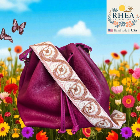 Purple handbag with a decorative purse strap in a field of flowers and butterflies, featuring the RHEA brand.