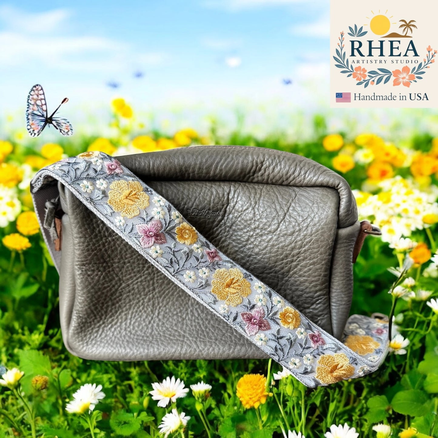 Gray leather bag with floral purse strap on a grassy field with flowers, RHEA brand visible