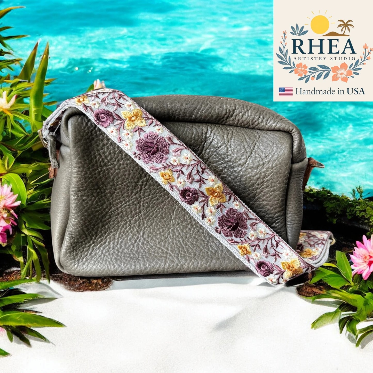 Gray leather bag with a floral purse strap against a scenic background, featuring the RHEA Artistry Studio logo.