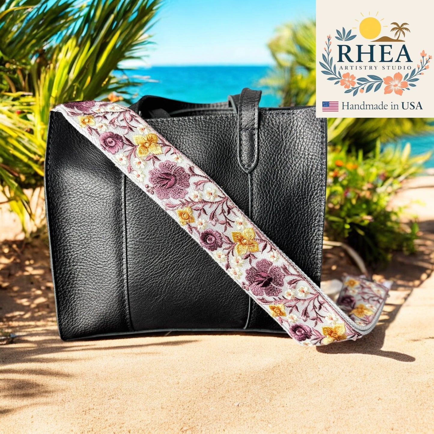 Black handbag with a floral purse strap on a beach, featuring the RHEA Artistry Studio logo.