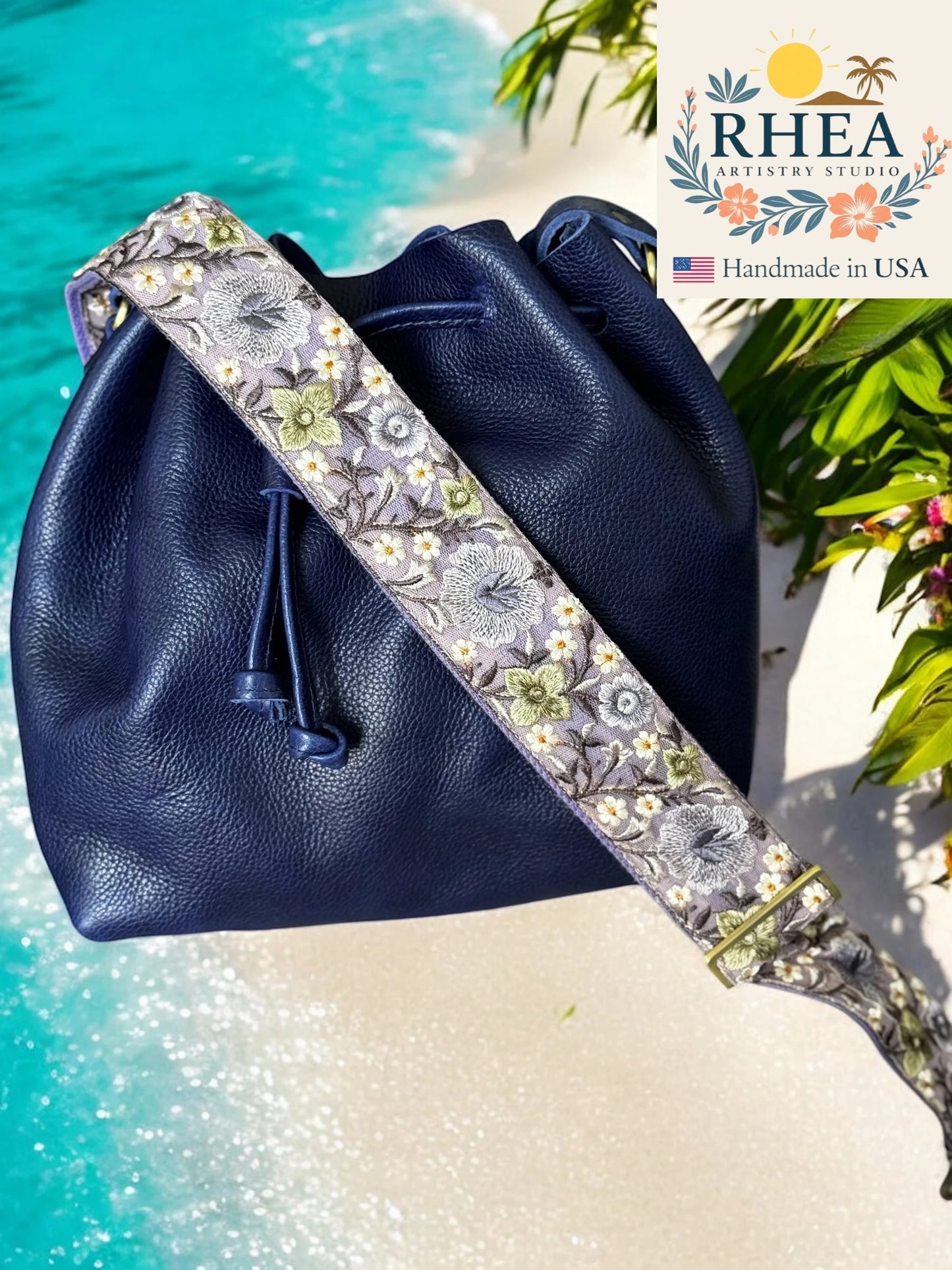 Navy blue leather bag with floral purse strap on a beach background, RHEA brand visible.